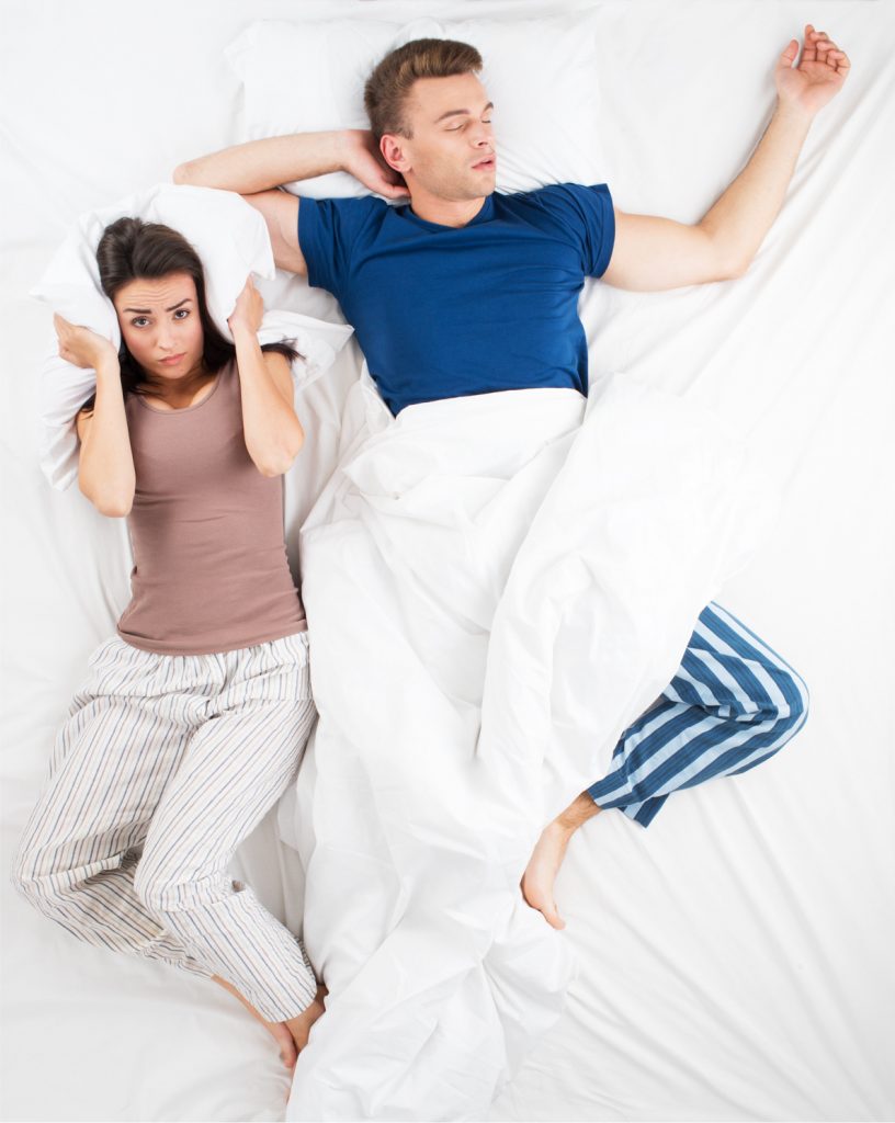 Couple in bed, man on his back snoring with woman covering her ears with a pillow